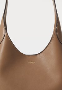 Sac à épaule en cuir marron avec bords supérieurs incurvés, détail de couture et petit logo doré « Coach New York » centré sous l’ouverture.