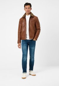 Bruine leren jas met een gestructureerde afwerking en een nepbont kraag, gecombineerd met blauwe jeans en crèmekleurige sneakers.