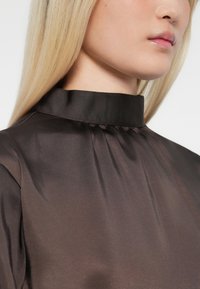 Close-up of a person wearing a dark brown satin high-neck top with slight gathers below the collar.