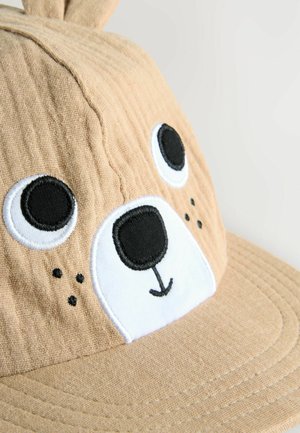 Cappello di tessuto beige con design di volto d'orso; presenta occhi, naso e bocca ricamati in bianco e nero; texture morbida e forma arrotondata.