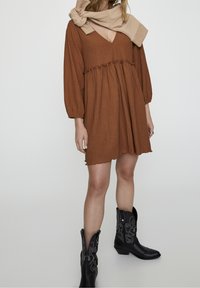 Rostfarbenes Kleid mit strukturiertem Stoff, gepufften Ärmeln und V-Ausschnitt. Kombiniert mit schwarzen Cowboy-Stiefeln mit dekorativen Nähten.