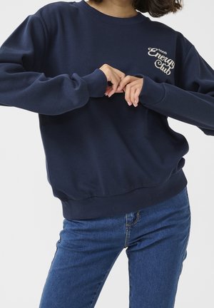 Osoba w granatowej bluzie z napisem „Good Energy Club” na piersi, ubrana w niebieskie dżinsy, z rękami złożonymi na talii.