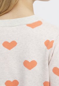 Pull gris clair à col rond, orné d'un motif répétitif de cœurs orange. Le tissu semble doux avec une texture confortable.