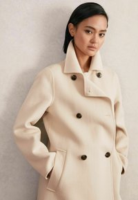 Beige wollen jas met een dubbelzijdig ontwerp, grote bruine knopen, brede kraag en zijzakken. Zachte textuur en getailleerde pasvorm.