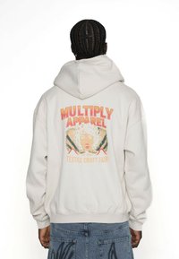 Bílá mikina s kapucí s potiskem na zádech, který zobrazuje boldní text "MULTIPLY APPAREL" nad obrázkem ženy, v pastelových tónech a uvolněném střihu.