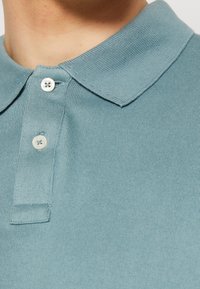 Tyrkysové polo tričko s ribovanou texturou, s klasickým límečkem a dvěma bílými knoflíky u zapínání. Detailní pohled se zaměřuje na výstřih.