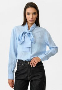 Lichtblauwe blouse met lange mouwen en een grote strik bij de kraag, gemaakt van gestructureerde stof, gecombineerd met donkere spijkerbroek.