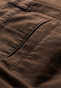 Hnědá herringbone tkanina s podrobnou kapsou s texturovaným povrchem a viditelným šitím podél okrajů.