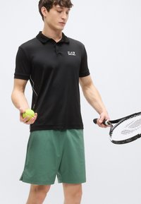 Juoda polo marškinėliai trumpomis rankovėmis, su apykakle ir baltais akcentais. Derinami su žaliais sportiniais šortais. Laiko žalią teniso kamuoliuką ir raketę.