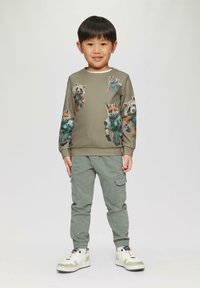 Olivgrüner Hoodie mit bedruckten Cartoon-Tieren in Raumanzügen. Kombiniert mit hellgrünen Cargo-Hosen und weißen Sneakers.