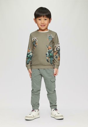 Olivgrüner Hoodie mit bedruckten Cartoon-Tieren in Raumanzügen. Kombiniert mit hellgrünen Cargo-Hosen und weißen Sneakers.