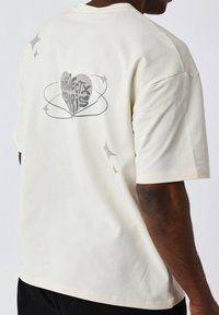 Camiseta de algodón color blanco roto con un corte relajado, que presenta un gráfico de corazón que dice "Protege París" rodeado de acentos de estrellas en la parte posterior.