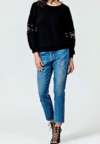 Maglione nero con dettagli in pizzo e perline sulle maniche, abbinato a jeans a gamba dritta blu chiaro e tacchi neri con lacci.