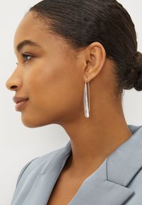 Boucles d'oreilles allongées en argent avec un design courbé et une finition polie, portées par une personne sur un fond neutre.