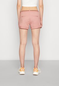 Light pink shorts with rolled cuffs, featuring a braided waistband and two back pockets. Paired with white sneakers having orange accents.