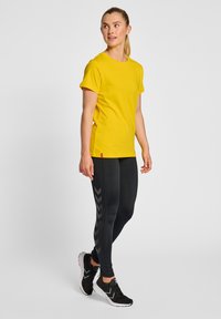 Geel T-shirt met korte mouwen gecombineerd met zwarte leggings met een grijs chevronpatroon. Zwarte sportschoenen maken de outfit compleet.