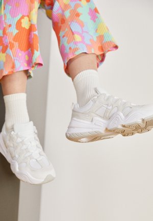 Person mit weißen Sneakers, weißen Socken und bunter Blumenhose sitzt mit baumelnden Beinen vor einem schlichten Hintergrund.