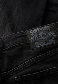 Czarny denim z teksturowaną skórzaną łatą z napisem "Superdry Standard Denim Goods" w szarym tłoczonym napisie.