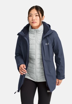 Donna che indossa una giacca con cappuccio blu navy sopra una giacca trapuntata grigia chiara con zip e pantaloni neri, mentre sistema la zip della giacca.
