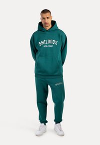 Conjunto de sudadera y pantalones de chándal en color verde azulado, fabricado con un tejido suave. La sudadera cuenta con un bolsillo frontal y el logo de la marca; los pantalones tienen el logo a lo largo del lateral.