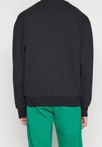 Sudadera negra con cuello redondo y mangas largas, con puños y dobladillo acanalados. Combinada con pantalones de chándal verdes.