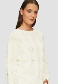 Maglione bianco lavorato a maglia con motivo a treccia, scollatura ampia e maniche a vestibilità larga. La texture sembra morbida e accogliente.