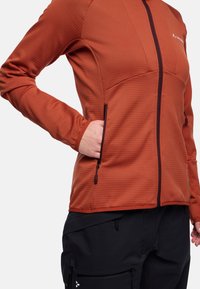 Giacca in pile color ruggine con zip frontale, tessuto testurizzato e tasche laterali. Abbinata a pantaloni neri con dettagli con zip.