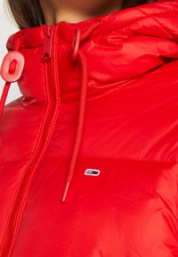 Giacca imbottita rossa con finitura lucida, dotata di chiusura a zip, cappuccio arricciato con coulisse regolabili e una piccola toppa con logo.