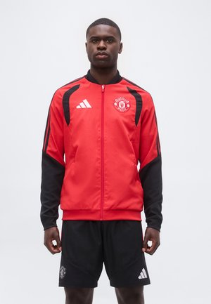 Giovane uomo con giacca con zip rossa e nera del Manchester United e pantaloncini neri con loghi, in piedi davanti a uno sfondo chiaro e uniforme.