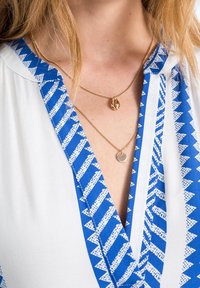 Des colliers en or superposés, l'un avec un pendentif en coquillage et l'autre avec une pièce texturée, portés sur un chemisier à motifs bleu et blanc avec un col en V.