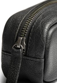 Detaljni prikaz ugla crne teksturirane kožne torbe s metalnim patentnim zatvaračem i kožnom trakom za povlačenje.