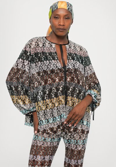Woman wearing a multicolored patterned knit top and matching pants with a colorful headscarf, standing against a plain background.
