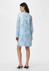 Femme portant une robe légère bleu clair avec des motifs floraux brodés, debout de profil avec des sandales à talons noires sur fond blanc.