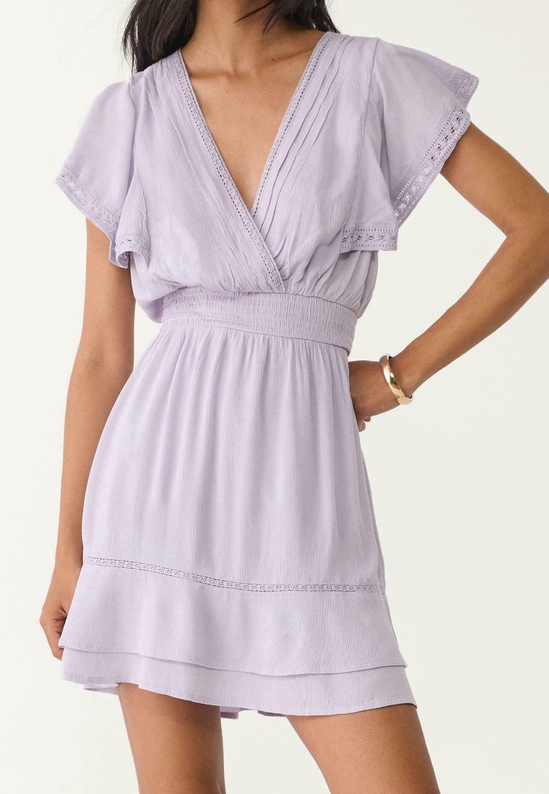 Femme portant une robe courte à manches courtes violet clair avec décolleté en V, taille smockée, jupe à volants et bordure en dentelle, avec un bracelet en or à son poignet.