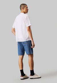Camisa blanca de manga corta, combinada con pantalones cortos de mezclilla azules y zapatillas ligeras. Presenta un ajuste holgado y una textura lisa.