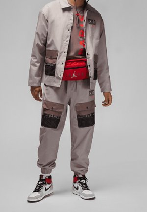 Mand iført lysegrå cargobukser og jakke med sorte mesh-lommer, rød hoftepose, grå T-shirt med rødt print og hvide, sorte-røde sneakers.