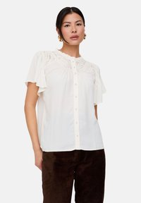White short-sleeve blouse with lace detail and ruffle trim, featuring a buttoned front, worn with dark brown pants. Smooth texture.