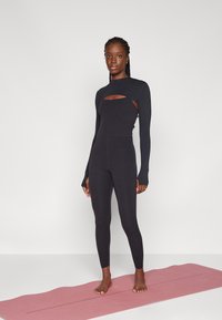 Must värvi pikkade varrukatega jumpsuit, mille rinnal on lõike detail ja keha lähedal jalgade lõpp, valmistatud venivast kangast, mis on asetatud roosale joogatamatile.