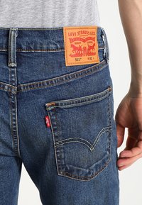 Levi's 511 farkut sinisessä denimmateriaalissa, joissa on oranssi nahkattu etiketti brändin logolla, kontrastiväriset ompeleet ja näkyvä punainen tarra takapussissa.