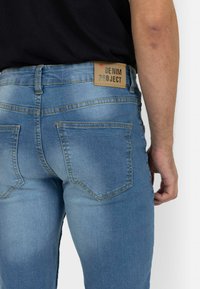 Svijetloplave traperice srednjeg struka, s pojasnim džepovima i kožnom etiketom na pojasu s natpisom "DENIM PROJECT".