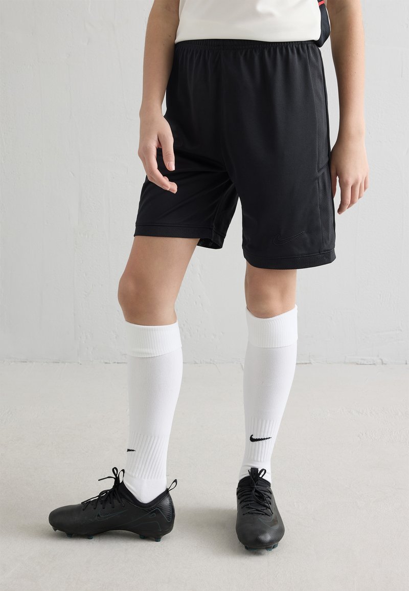 Pessoa vestindo calções pretos de futebol Nike, meias altas brancas da Nike e chuteiras pretas da Nike, em pé num chão claro com fundo de parede branca.