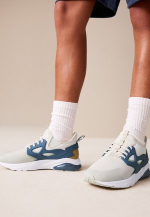 Nogi w białych skarpetkach i jasnoszarych tenisówkach z niebieskimi i brązowymi akcentami, stojące na jasnej powierzchni na neutralnym tle.