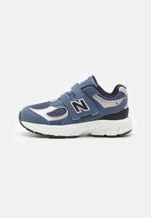 Blå og hvide New Balance sneakers med velcrobånd, sort inderside og en tyk hvid sål, vist fra siden på en ensfarvet baggrund.