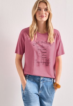 Femme aux longs cheveux blonds portant un t-shirt rose avec une illustration de café et un jean bleu, debout les mains dans les poches.