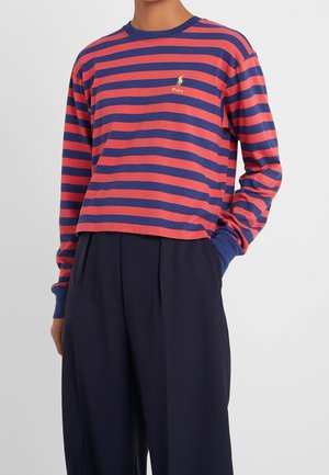 Person trägt ein langärmliges Hemd mit roten und marineblauen horizontalen Streifen, einem gelben Polo-Logo und marineblaue, hoch taillierte Hose.