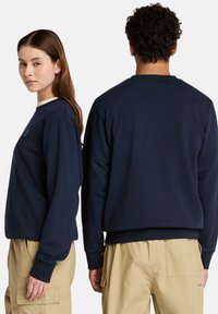 Felpa blu navy con collo a giro, polsini e orlo a costine. Presenta un piccolo logo ricamato sul davanti. Indossata con pantaloni cargo beige.