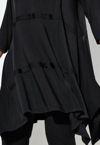 Vapaa musta tunika epäsymmetrisellä helmalla ja satiininauhadetaljeilla, päällä tiukasti istuvien mustien housujen.