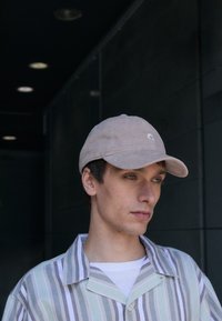 Căciulă bej din corduroy cu o margine curbată și un detaliu subtil al logo-ului. Modelul poartă o cămașă cu mânecă scurtă și dungi în culori deschise. Fundalul este întunecat.