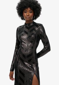 Vestido negro de lentejuelas con mangas largas, cuello simulado y patrón de chevrón. Presenta una abertura alta en el muslo para un detalle adicional. Textura brillante en todo el vestido.