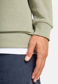 Olijfgroen katoenen sweatshirt met geribbelde manchetten, gelaagd over een wit shirt en donkerblauwe ribfluwelen broek, met een hand die op de stof rust.